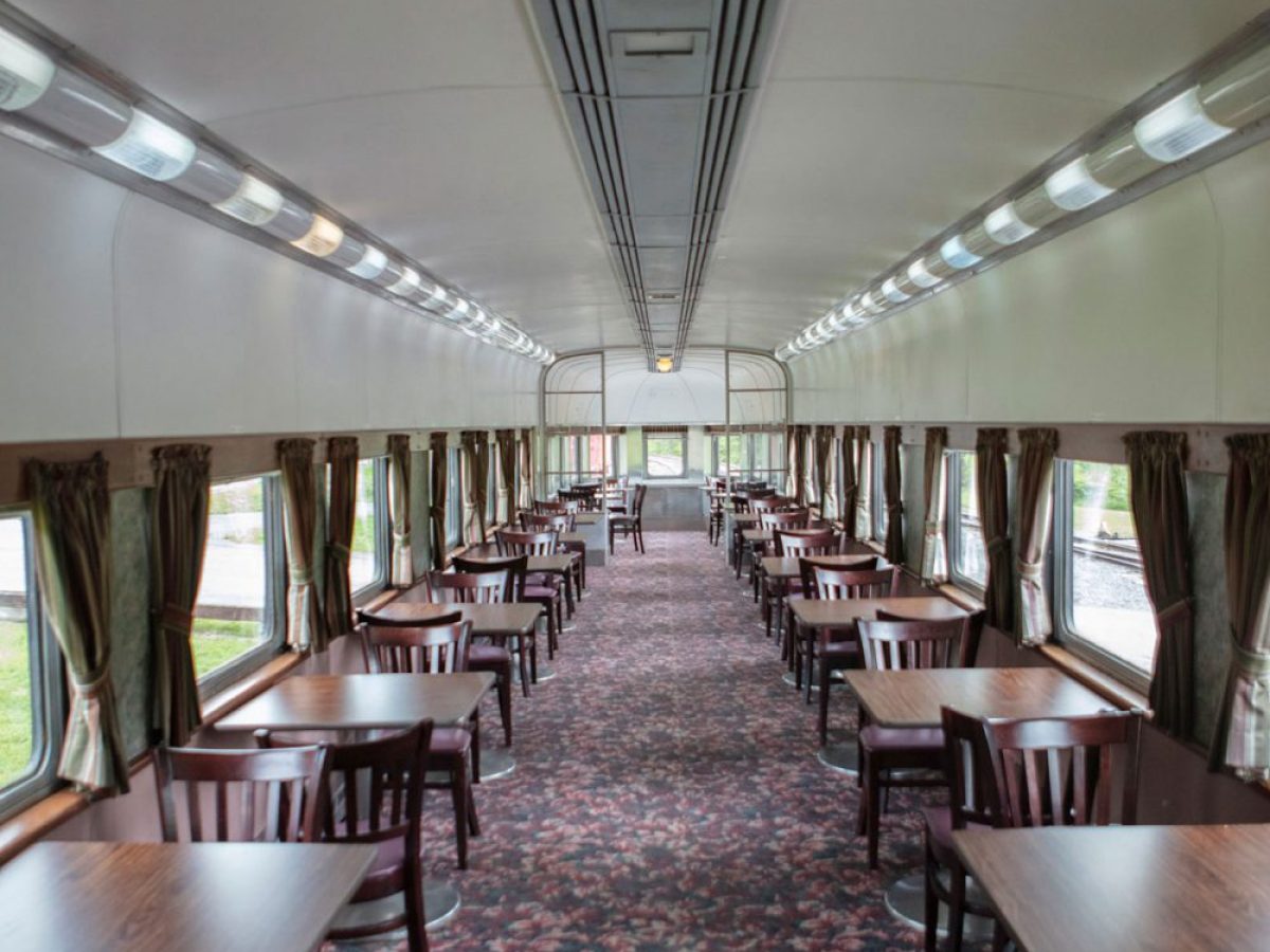 Interior of an empty train dining car with wooden tables and chairs, lined alongside windows.
