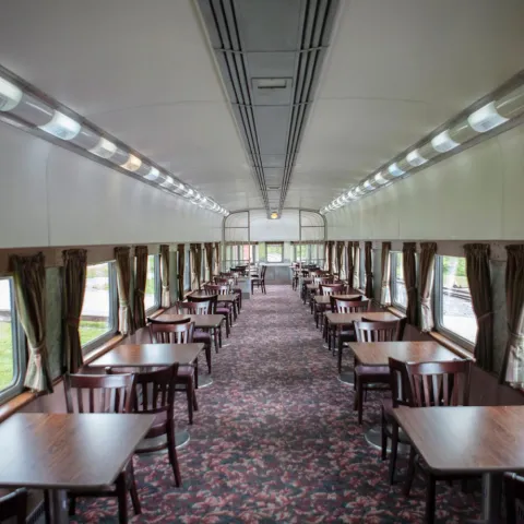 Interior of an empty train dining car with wooden tables and chairs, lined alongside windows.