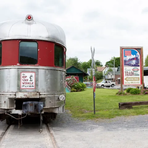 Vintage train car with 'Rip Van Winkle Flyer' sign and nearby bus, buildings, and train ride sign.