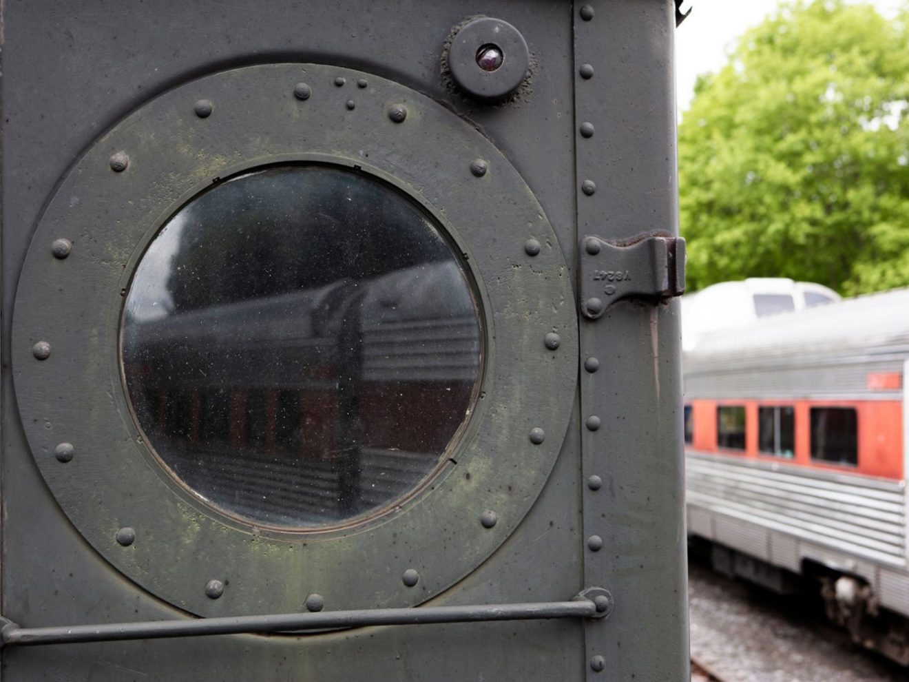 Round window on vintage train door with another train visible outside.