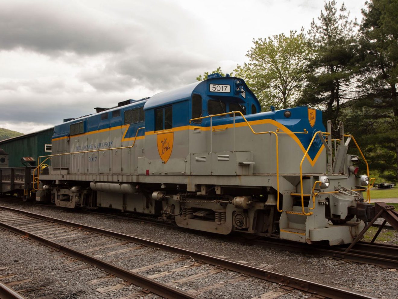 Blue and gray locomotive parked on railway tracks with trees and cloudy sky in the background.