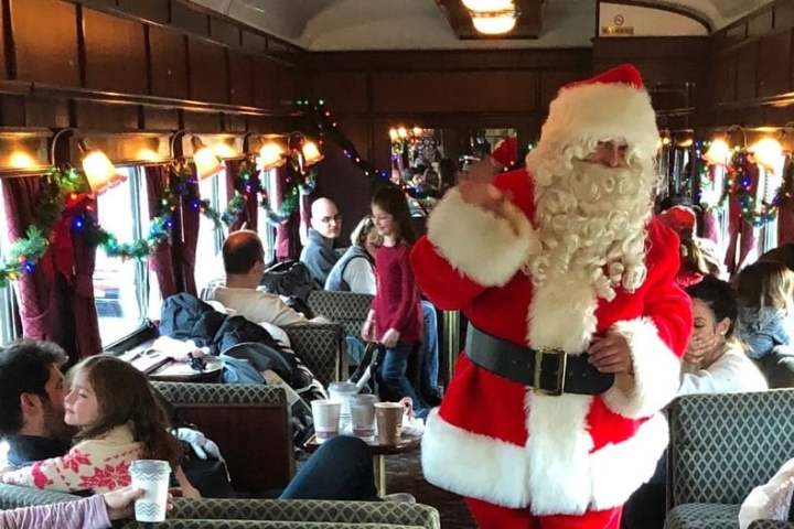 Santa Claus walking through a decorated train car with seated passengers.